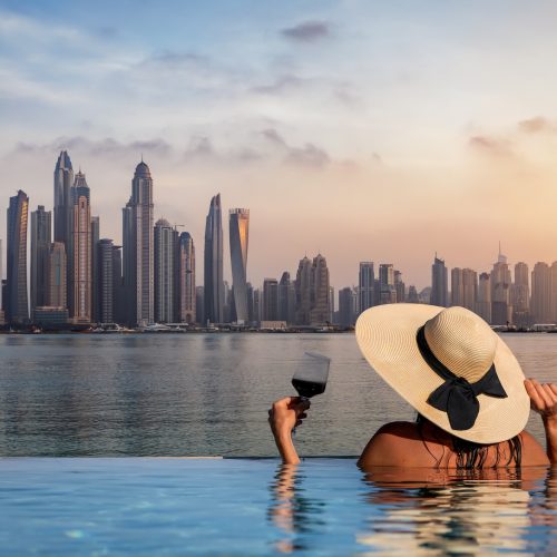 A elegant woman with a hat and a drink in her hand stands at the edge of a infinity pool and enjoys the view to the skyline of the Dubai Marina during sunset, UAE